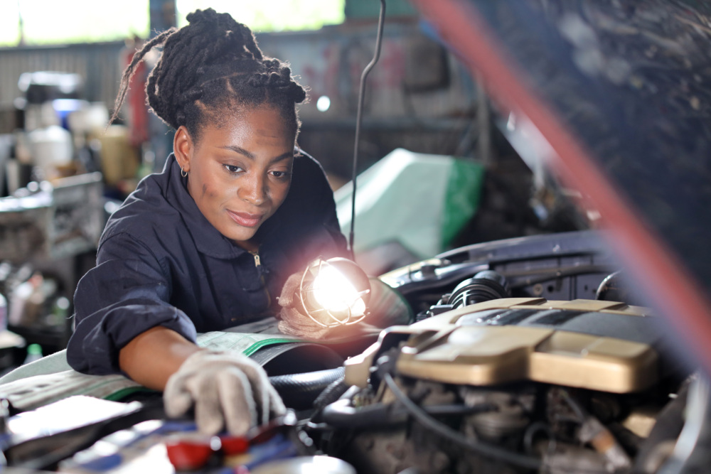 Woman in a workshop holding a light, inspecting a car engine.